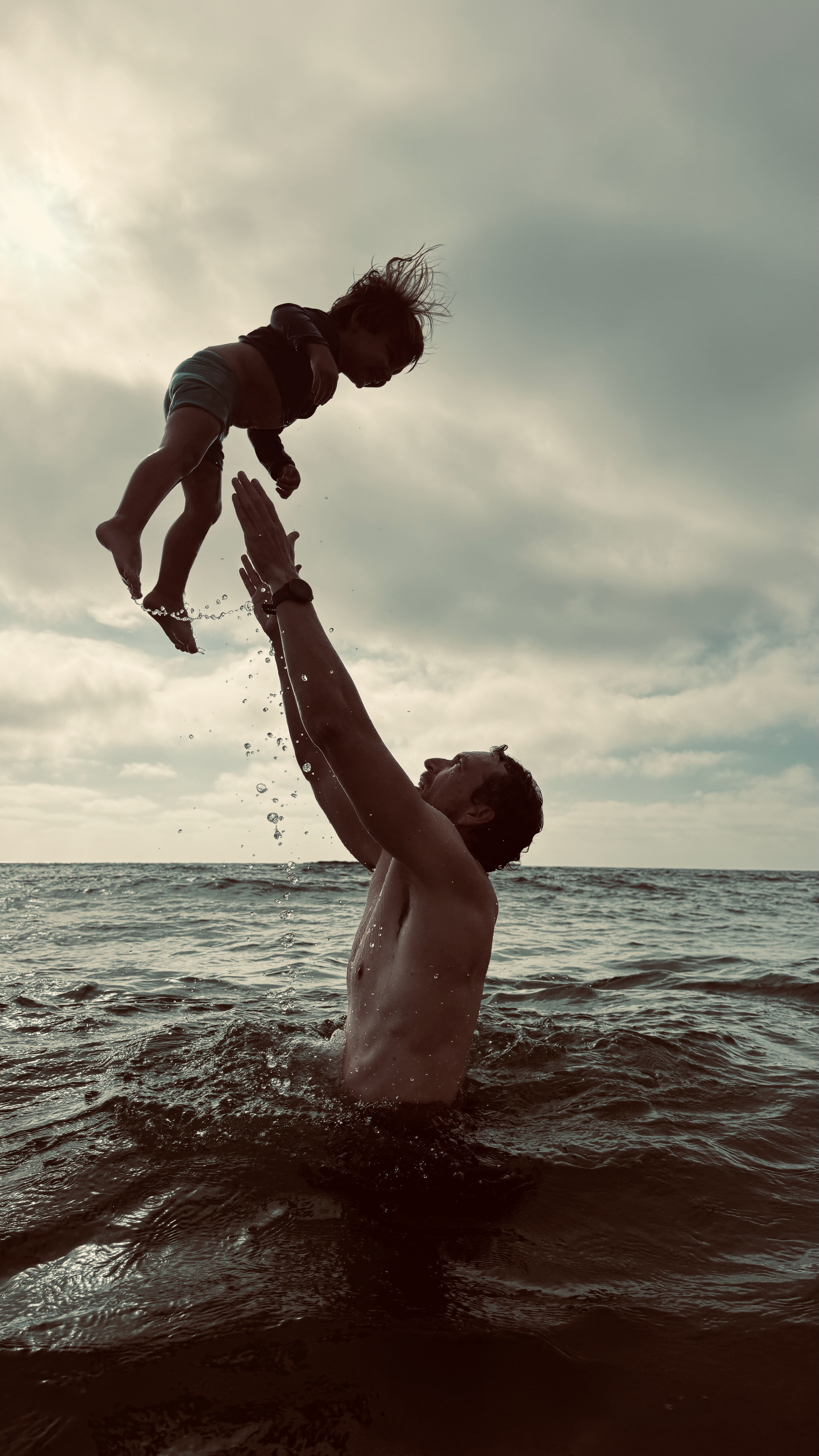 A parent lifting a small child joyfully above the sea — both silhouetted against the sky.